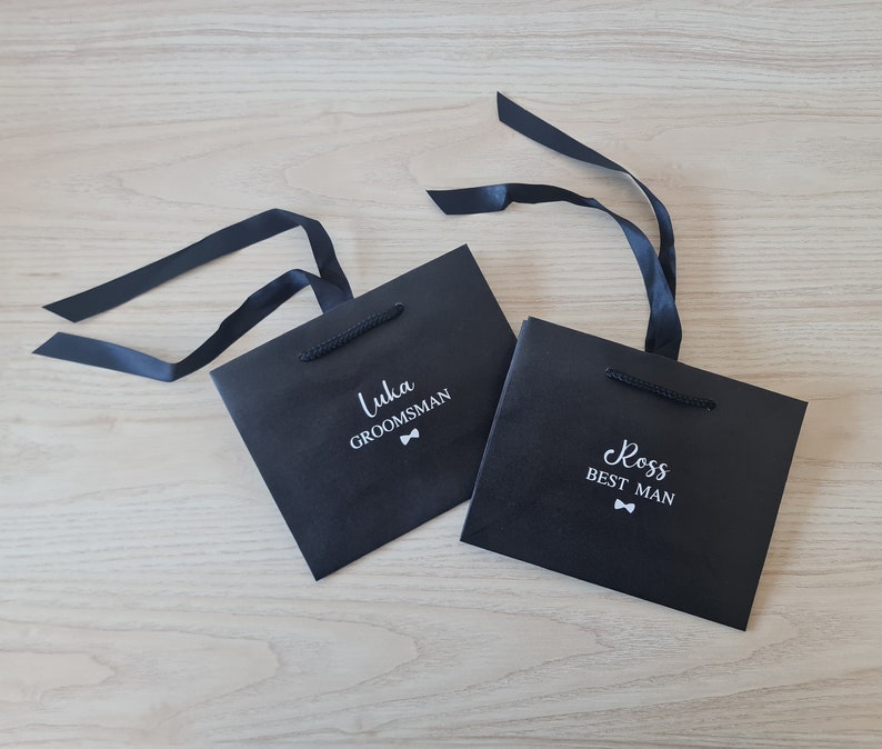 Pode incluir: Duas sacolas de presente pretas com texto branco e um s&iacute;mbolo de gravata borboleta. A sacola &agrave; esquerda diz "Luka Groomsman" e a sacola &agrave; direita diz "Ross Best Man".
