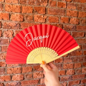May include: A red folding hand fan with a light-colored wooden frame featuring intricate cut-out patterns. The fabric portion of the fan displays the name Jacqui written in a white cursive script font across the upper left side of the red surface.
