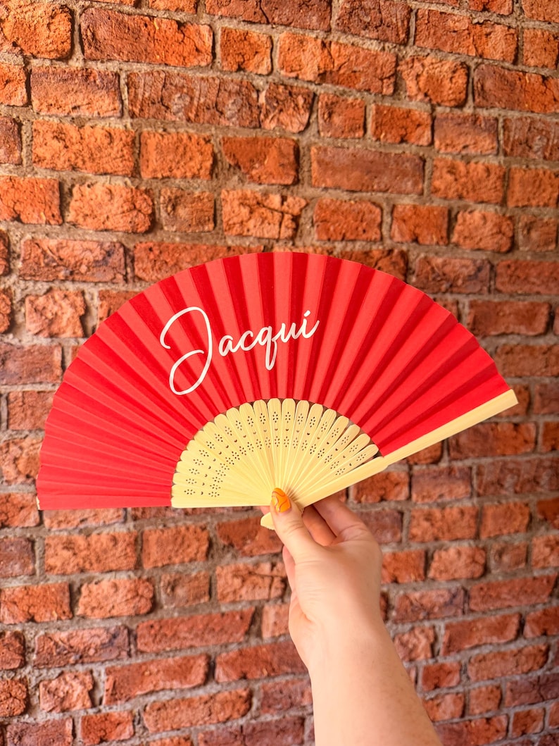 May include: A red folding hand fan with a light-colored wooden frame featuring intricate cut-out patterns. The fabric portion of the fan displays the name Jacqui written in a white cursive script font across the upper left side of the red surface.