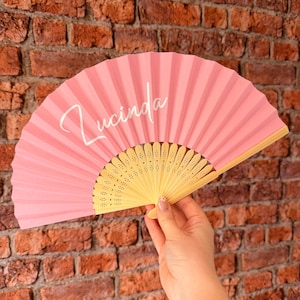 May include: A handheld folding fan with a solid pink fabric canopy and a light-colored wooden frame. The name Lucinda is printed in white cursive script across the left side of the pink fabric. The fan features a series of small, decorative cutouts.