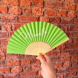 May include: A lime green folding hand fan featuring the name Michelle printed in white script lettering across the center. The fan has a light-colored wooden frame with intricate cut-out patterns at the base and pleated fabric panels.