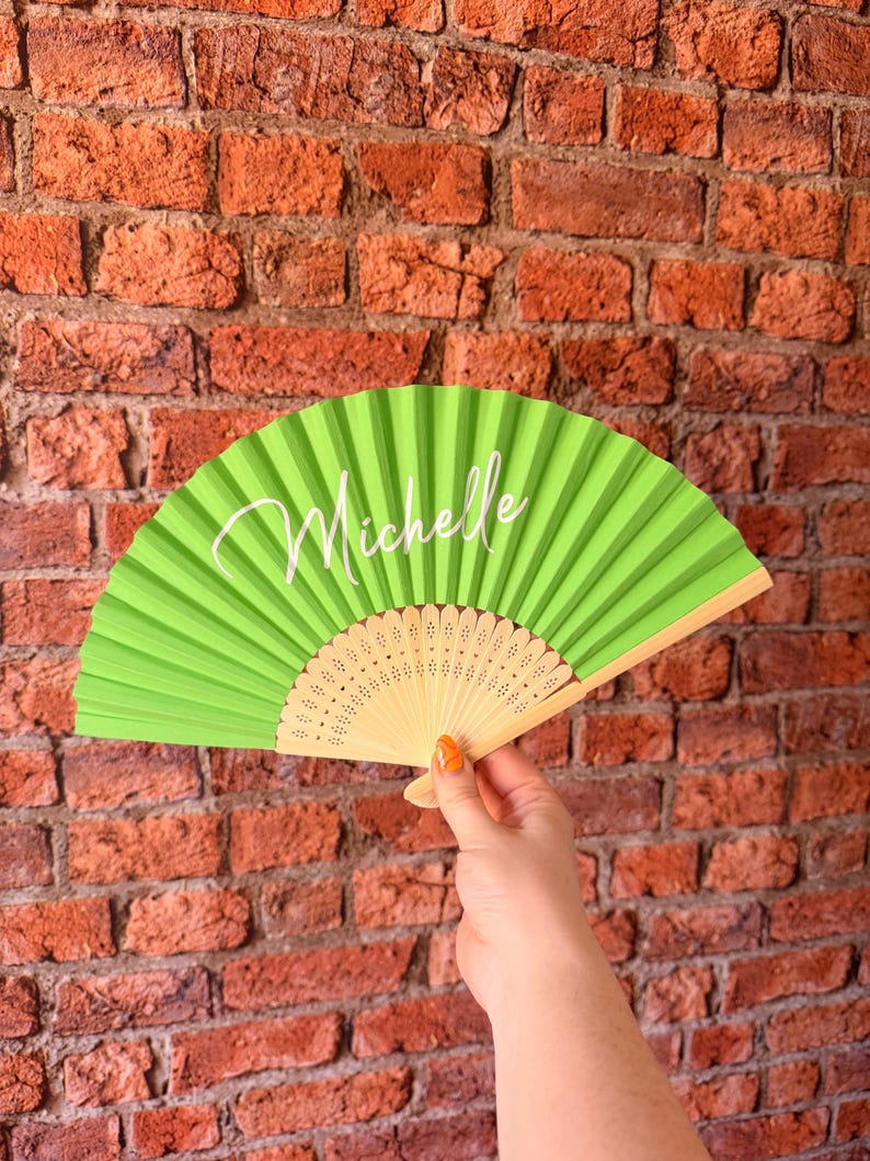 May include: A lime green folding hand fan featuring the name Michelle printed in white script lettering across the center. The fan has a light-colored wooden frame with intricate cut-out patterns at the base and pleated fabric panels.