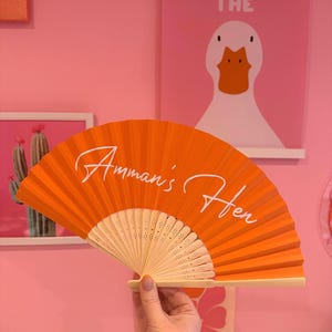 May include: A bright orange folding hand fan with a light-colored wooden frame. The fan features the white cursive text "Amman's Hen" printed across the center of the pleated fabric surface. The fan is held open to display the full design.