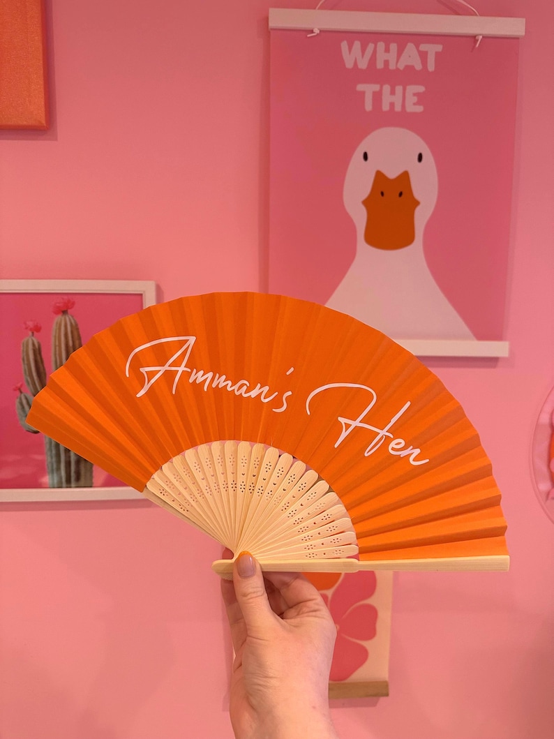 May include: A bright orange folding hand fan with a light-colored wooden frame. The fan features the white cursive text "Amman's Hen" printed across the center of the pleated fabric surface. The fan is held open to display the full design.