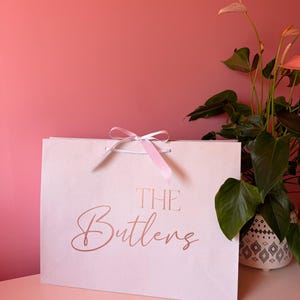May include: A cream gift bag with the words "THE Butlers" in rose gold script. The bag is tied with a pink ribbon. A potted plant with green leaves and pink flowers is to the right of the bag, against a pink wall.