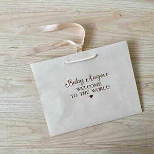 May include: A cream-colored gift bag with the text "Baby Angove WELCOME TO THE WORLD" in dark brown script. A small red heart is below the text. The bag has a cream-colored ribbon and a white rope handle.