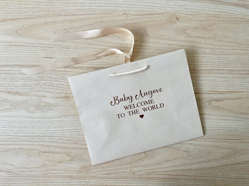 May include: A cream-colored gift bag with the text "Baby Angove WELCOME TO THE WORLD" in dark brown script. A small red heart is below the text. The bag has a cream-colored ribbon and a white rope handle.