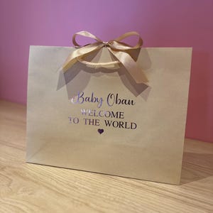 May include: A tan gift bag with a satin ribbon bow and handles. The bag has the text "Baby Oban WELCOME TO THE WORLD" printed in silver. A small purple heart is below the text. The bag is on a wooden surface.