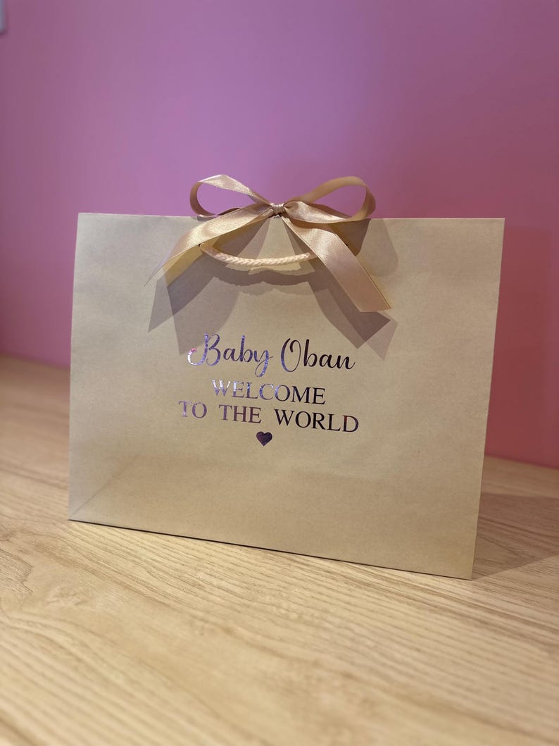 May include: A tan gift bag with a satin ribbon bow and handles. The bag has the text "Baby Oban WELCOME TO THE WORLD" printed in silver. A small purple heart is below the text. The bag is on a wooden surface.