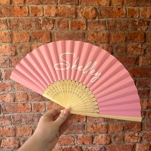 May include: A handheld folding fan with a solid pink fabric leaf and a light-colored wooden frame. The name Shelley is printed in a white cursive script across the center of the pink fabric. The wooden ribs feature small decorative cutouts.