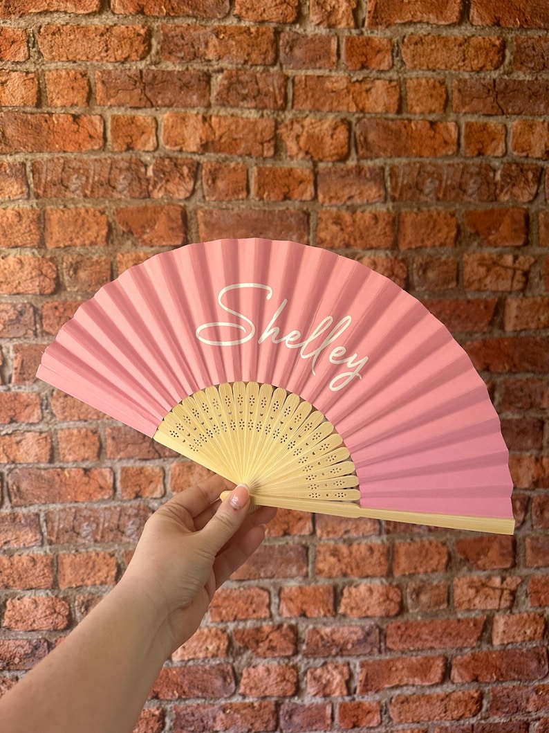 May include: A handheld folding fan with a solid pink fabric leaf and a light-colored wooden frame. The name Shelley is printed in a white cursive script across the center of the pink fabric. The wooden ribs feature small decorative cutouts.