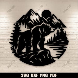 Peut inclure: Décoration murale en métal noir représentant un ours sur un rocher, avec des montagnes, des arbres et un soleil en arrière-plan. Le design circulaire comprend le texte "SVG DXF PNG PDF" en bas.