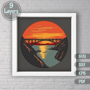 May include: A 9-layer paper cut design of a bridge over a body of water with a sunset in the background. The design is in a circular frame.