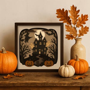 May include: A framed Halloween-themed shadow box featuring a haunted house, witch, bats, and ghosts. Two carved pumpkins and a white pumpkin sit on a wooden surface, with a vase of autumn leaves to the right.