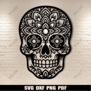 Puede incluir: Calavera negra con intrincados diseños florales, cortada de una sola pieza. La calavera presenta cuencas de ojos detalladas, una nariz y una dentadura completa. El diseño incluye el texto "SVG DXF PNG PDF" en la parte inferior. La calavera está sobre una pared de ladrillos blancos.
