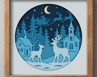 May include: A framed paper cut artwork of a winter scene. The layered design uses a blue colour palette, showing a church, houses, deer, and Santa's sleigh under a crescent moon and stars. The frame is made of wood.