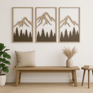 May include: Three framed wall art pieces with a mountain and forest design. The artwork features a light brown mountain silhouette above a dark brown forest. The frames are light brown. A bench, pillow, and side table are in the foreground.