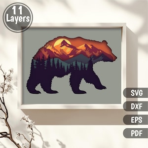 May include: A framed artwork featuring a layered design of a bear silhouette with a mountain and forest scene. The artwork is predominantly dark purple, with orange, yellow, and green accents. The text "11 Layers" is in a circle in the upper left corner. The image also includes the file types: SVG, DXF, EPS, and PDF.
