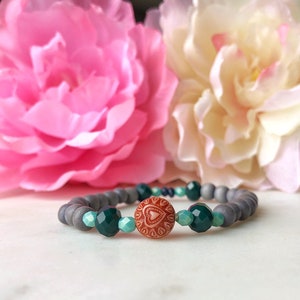Peut inclure: Un bracelet en perles grises avec des perles de verre vertes et bleues et un bouton en forme de cœur avec un motif floral.