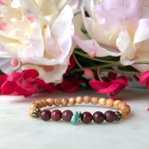 May include: A wooden bracelet with red and brown beads and a turquoise stone accent. The bracelet is on a white surface with pink and red flowers in the background.
