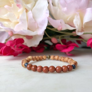 Peut inclure: Un bracelet en perles brunes et beiges avec deux perles bleu foncé. Le bracelet est sur une surface blanche avec des fleurs roses en arrière-plan.
