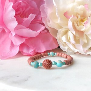 May include: A wooden bracelet with turquoise and brown heart-shaped beads. The bracelet is on a white surface with pink and white flowers in the background.