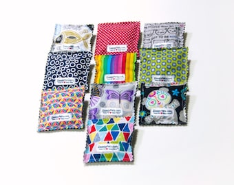 May include: Nine colorful, patterned fabric squares, likely cat toys, are arranged on a white surface. Designs include fish, polka dots, rainbows, butterflies, and geometric shapes. Each square has a tag with text.