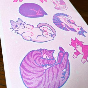 May include: A pink and purple print with six illustrations of cats in various poses. The cats are drawn in a cartoon style with simple lines and rounded features. The background is a light pink with a subtle sparkle effect.