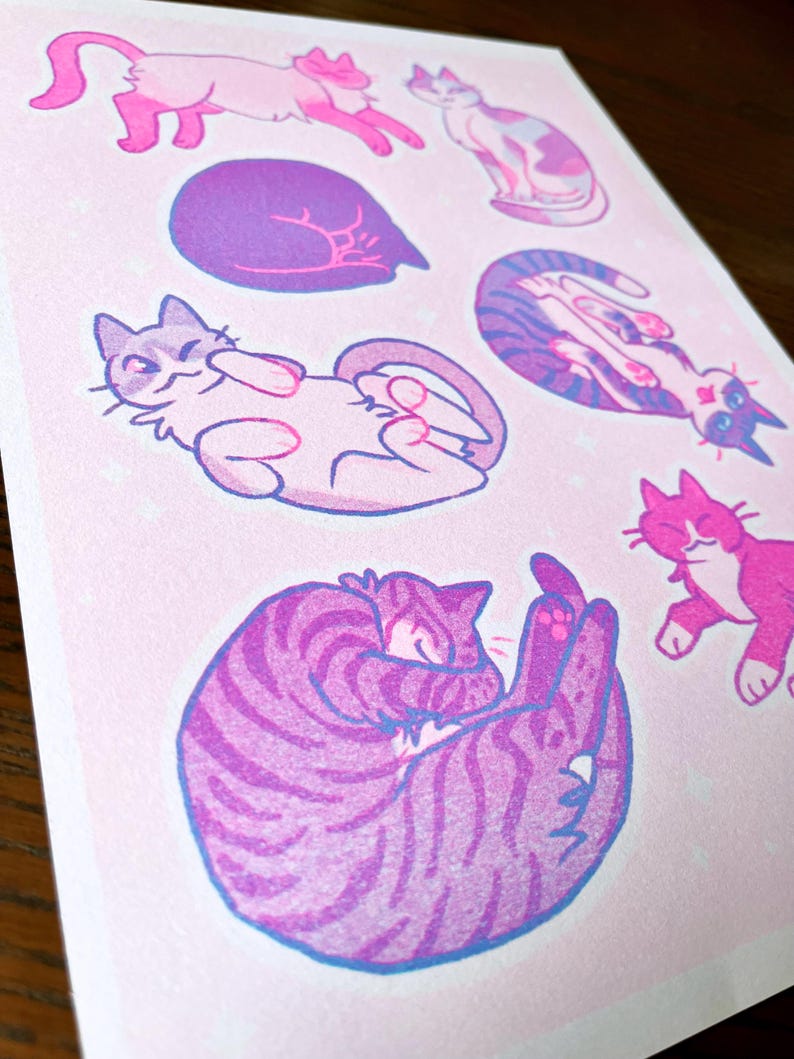 May include: A pink and purple print with six illustrations of cats in various poses. The cats are drawn in a cartoon style with simple lines and rounded features. The background is a light pink with a subtle sparkle effect.