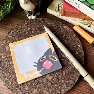 Könnte beinhalten: Ein gelber Haftnotizblock mit einer schwarzen Katzenillustration auf der rechten Seite. Die Katze hat große, runde Augen und einen offenen Mund mit einer rosa Zunge. Der Notizblock liegt auf einem Korkuntersetzer mit einem Stift und einem Textmarker daneben.