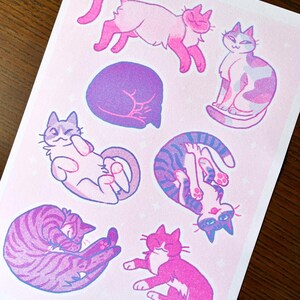 May include: A set of six illustrated stickers featuring cats in various poses. The stickers are printed on a pink background with white stars. The cats are drawn in a cartoon style and are colored in shades of pink, purple, and blue.
