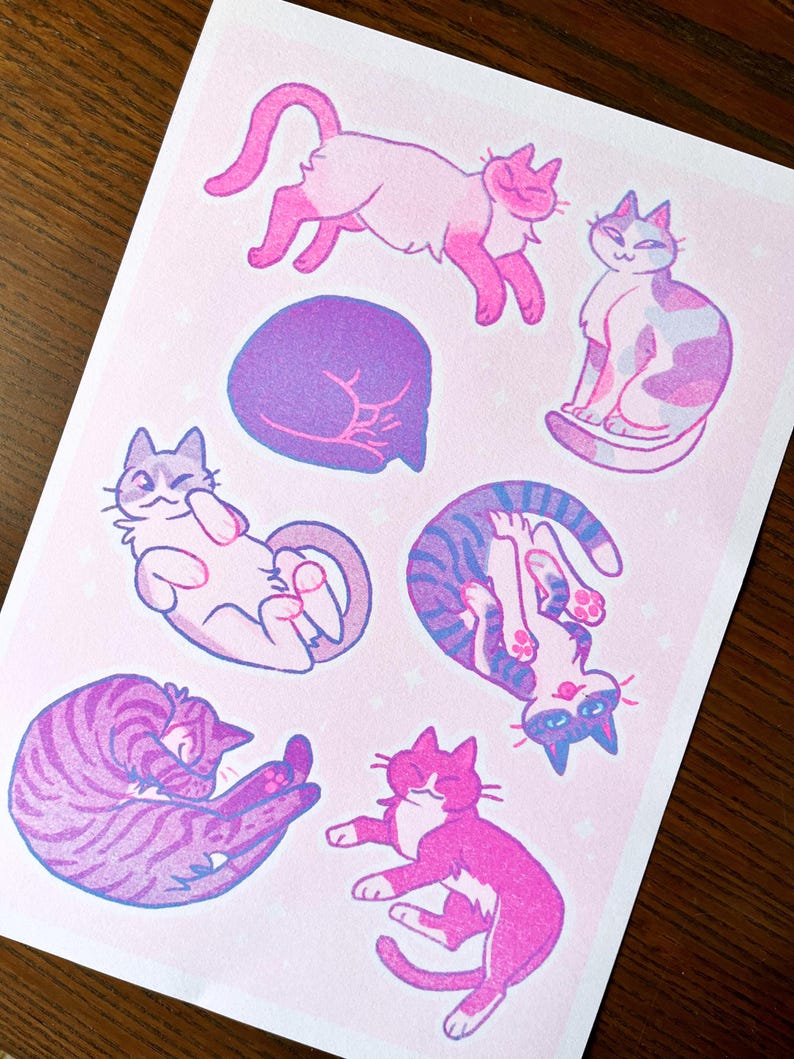 May include: A set of six illustrated stickers featuring cats in various poses. The stickers are printed on a pink background with white stars. The cats are drawn in a cartoon style and are colored in shades of pink, purple, and blue.