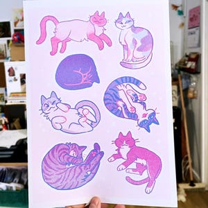 May include: A print featuring nine illustrations of cats in various poses. The cats are drawn in a cartoon style with pink, blue, and purple colors. The background is a light pink with a subtle sparkle effect.