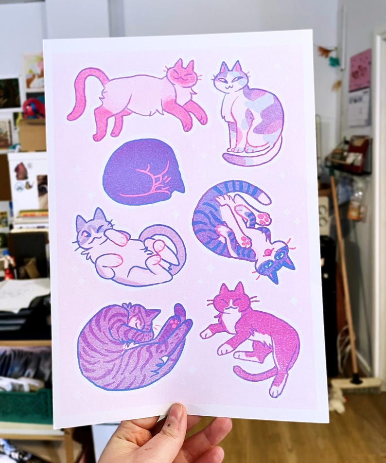 May include: A print featuring nine illustrations of cats in various poses. The cats are drawn in a cartoon style with pink, blue, and purple colors. The background is a light pink with a subtle sparkle effect.