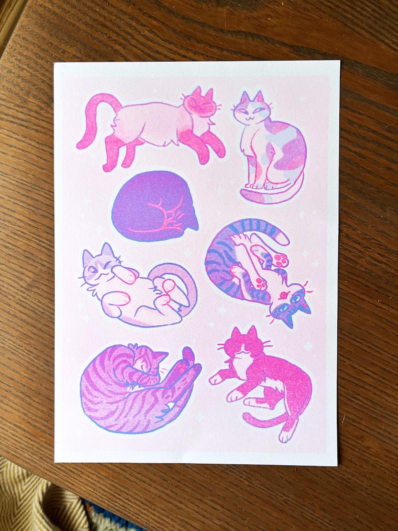 May include: A pink and purple print featuring seven cats in various poses. The cats are drawn in a cartoon style with simple lines and bright colors. The background is a light pink with a subtle sparkle effect.