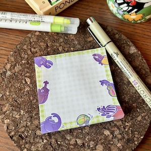 Può includere: Un blocco note verde e viola con un motivo a scacchi e illustrazioni di scarabei dei cartoni animati attorno al bordo. Il blocco note è su una superficie di sughero.