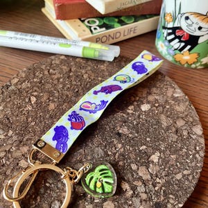 May include: A yellow and white keychain with a gold key ring and a green leaf charm. The keychain features a ribbon with a repeating pattern of purple, blue, and yellow bugs.