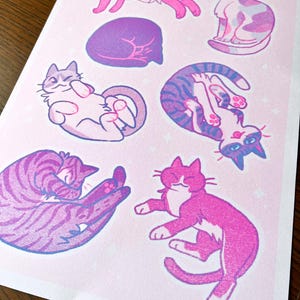 May include: A pink and purple print featuring nine different illustrations of cats in various poses. The cats are drawn in a cartoon style with simple lines and bright colors.
