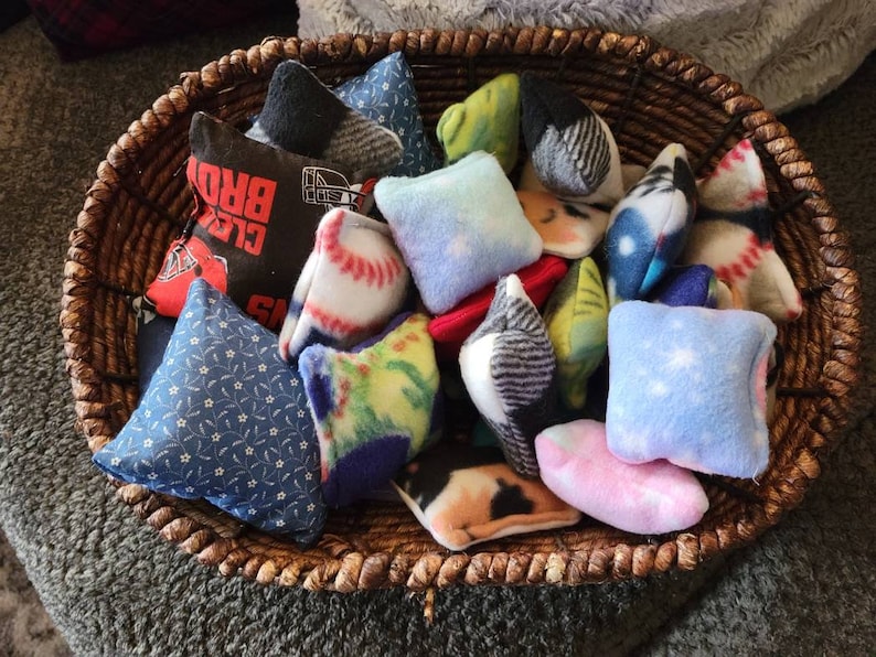 May include: A wicker basket filled with a variety of colorful fabric bean bags. The bean bags have different patterns, including baseballs, stars, and floral designs.
