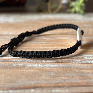 Op de afbeelding: Een zwart macram&eacute; armband met een zilveren accent. De armband is verstelbaar met een schuifknoopsluiting. Het macram&eacute; is strak geweven en het zilveren accent is een kleine, gladde rechthoek. De armband wordt getoond op een lichtbruin houten oppervlak.