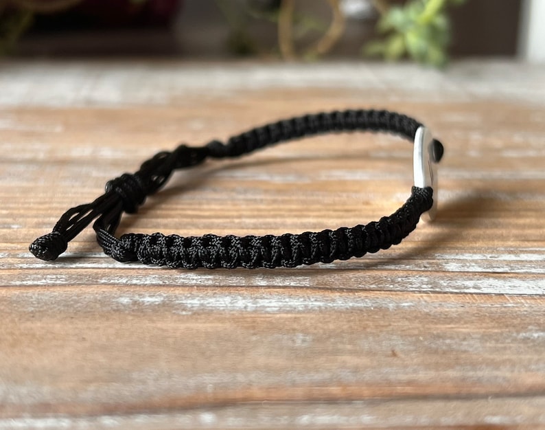 Op de afbeelding: Een zwart macram&eacute; armband met een zilveren accent. De armband is verstelbaar met een schuifknoopsluiting. Het macram&eacute; is strak geweven en het zilveren accent is een kleine, gladde rechthoek. De armband wordt getoond op een lichtbruin houten oppervlak.