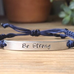 May include: A navy blue braided bracelet with a silver charm that reads "Be Strong".
