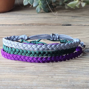 May include: A set of three braided friendship bracelets in purple, green, and gray.
