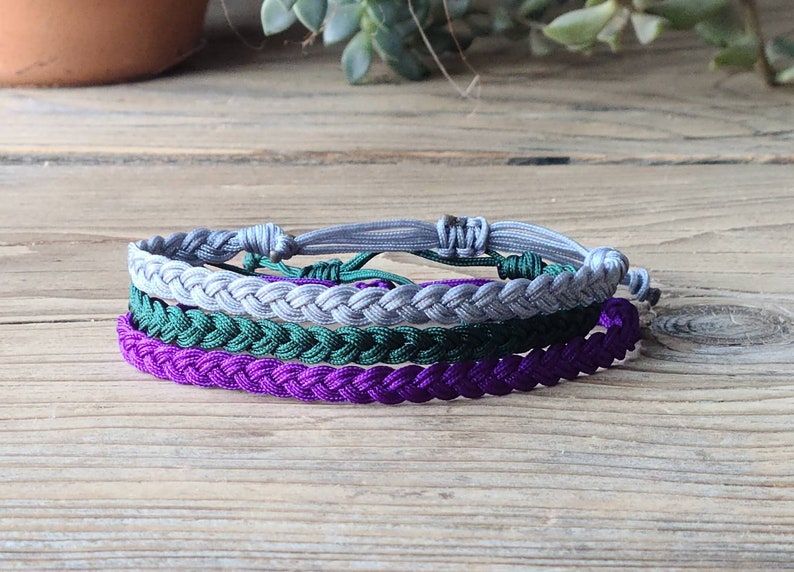 May include: A set of three braided friendship bracelets in purple, green, and gray.