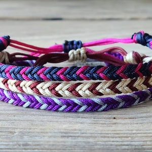 May include: Three braided friendship bracelets in purple, pink, and brown with white accents. The bracelets are woven in a chevron pattern.