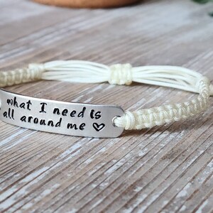 Puede incluir: Una pulsera trenzada blanca con un dije plateado que dice "what I need is all around me" con un s&iacute;mbolo de coraz&oacute;n.