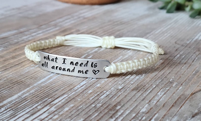 Puede incluir: Una pulsera trenzada blanca con un dije plateado que dice "what I need is all around me" con un s&iacute;mbolo de coraz&oacute;n.