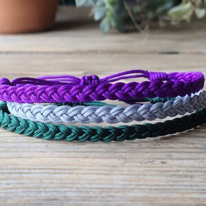 May include: Three braided bracelets in purple, gray, and green colors. The bracelets are stacked on top of each other.