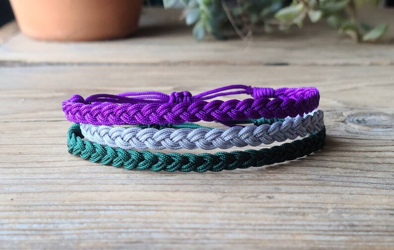May include: Three braided bracelets in purple, gray, and green colors. The bracelets are stacked on top of each other.