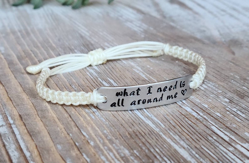 Puede incluir: Pulsera de cuerda blanca con un dije plateado que dice "what I need is all around me" con un s&iacute;mbolo de coraz&oacute;n.